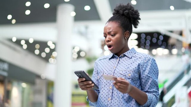 Shocked upset african american woman checks her credit card on mobile phone standing in mall shopping center. Frustrated female faced fraud, became victim of deception or glitch and error with banking
