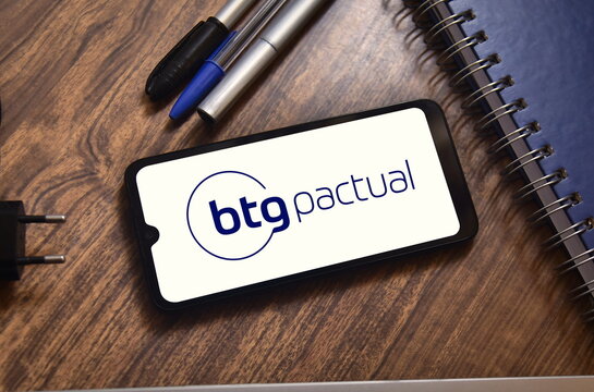 BTG Pactual logo on a cell phone screen over objects on a table. Brazilian investment bank, specializing in investment capital and venture capital, as well as investment fund management.