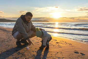 Toddler bends to sand while dad shows seashells. Family discoveries by the sea.