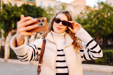 Cheerful woman in sunglasses blogs on phone while strolling along sunny city streets. Beautiful...