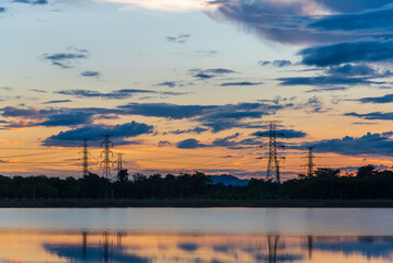Silhouette of high voltage electricity pylons transmission towers and power lines against a dramatic orange and blue sunset sky, reflected in a calm lake.