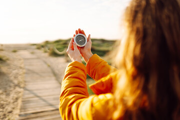 Fototapeta premium A young hiker in a bright jacket and backpack holds a compass and looks out at the seascape. Hiker chooses a direction while standing on a hiking trail at sunset. Concept of adventure, navigation.