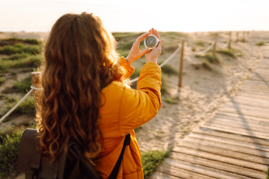 A young hiker in a bright jacket and backpack holds a compass and looks out at the seascape. Hiker chooses a direction while standing on a hiking trail at sunset. Concept of adventure, navigation.