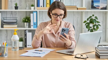 Young businesswoman wearing glasses, waving hand and using smartphone for video call while sitting at desk with laptop in modern office. Communication and remote work concept. - Powered by Adobe
