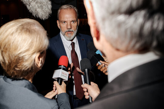 Middle aged Caucasian man speaking to group of journalists holding microphones during press conference, reporters interviewing and recording responses, news channel microphone visible