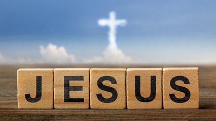 Row of wooden cubes with text of Jesus on the table with a cloud of Christian cross on a blue sky background. Concept of Christianity, religious, faith, Jesus, or belief
