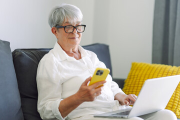 A modern elderly woman wearing glasses works on a laptop while sitting on a sofa, holding a phone. A cheerful older woman works on a laptop in a cozy room. Concept of modernity, technology.
