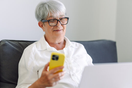 A modern elderly woman wearing glasses works on a laptop while sitting on a sofa, holding a phone. A cheerful older woman works on a laptop in a cozy room. Concept of modernity, technology. - Powered by Adobe