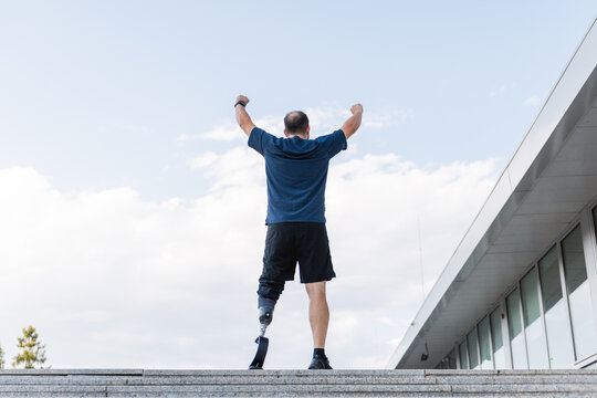 Man with a prosthetic leg raising arms in victory after successful running training up long street stairs. Disability, achievement, and motivation concept.