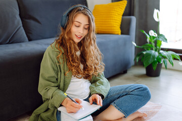 Obraz premium Young woman wearing wireless headphones sits in cozy living room and takes notes in notebook. Beautiful woman enjoys reading book or taking notes. Concept of education, relaxation, and comfort.