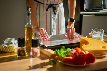 Woman in apron covering baking tray with foil surrounded by fresh vegetables, pumpkin and ingredients in kitchen with sunlight. Concept of home cooking and healthy meal preparation
