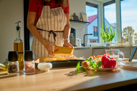 Woman in striped apron placing butternut squash or pumpkin on baking tray in bright kitchen with fresh vegetables and ingredients. Process of cooking at home and healthy food preparation - Powered by Adobe