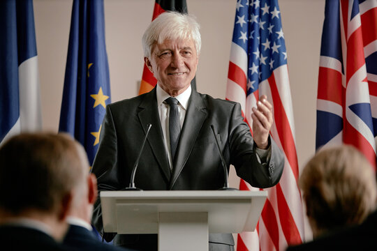 Senior Caucasian man speaking at podium gesturing with hand during press conference with international flags in background addressing audience of business professionals - Powered by Adobe