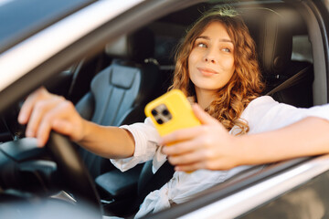 Portrait of a young woman driving with a phone. A beautiful woman driving a car in a busy parking lot, opening an app on smartphone. Concept: technology, city, lifestyle.
