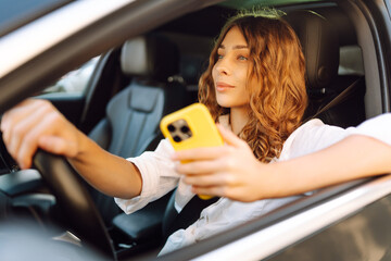 Portrait of a young woman driving with a phone. A beautiful woman driving a car in a busy parking lot, opening an app on smartphone. Concept: technology, city, lifestyle.