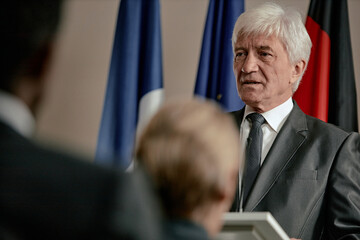 Caucasian senior man speaking in formal setting, standing in front of international flags,...
