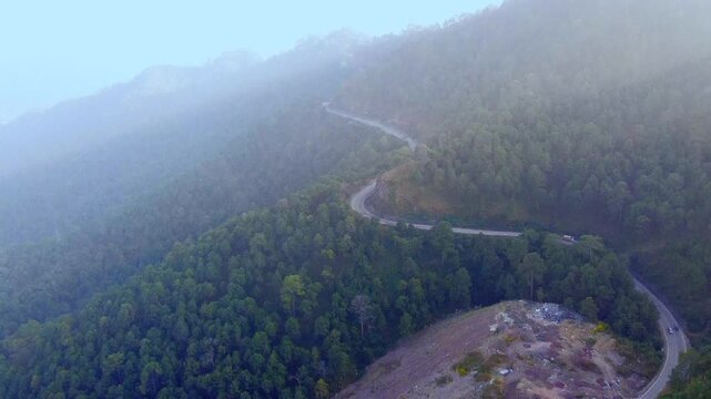 Aerial drone footage capturing the scenic NH-5 Chandigarh-Shimla Highway in the Himalayan foothills, features cars and trucks moving along the winding asphalt road, flanked by dense green forests. 