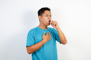 A young Asian man in a blue shirt coughs with his hand in front of his mouth and holds his chest, emphasizing the importance of hygiene and respiratory health.