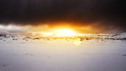 Winter landscape of snow-covered ground leading up to a rugged mountain range with dramatic sunlight through dark storm clouds. Natural background