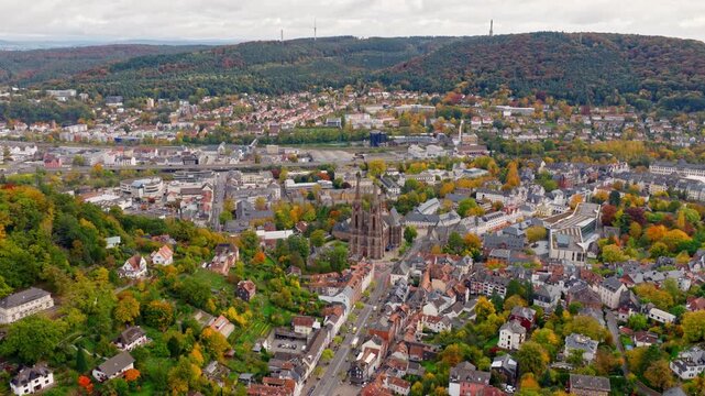 The drone flies over Marburg, showing the old town and the iconic Elisabeth Church, surrounded by trees and the forested hills of the region.