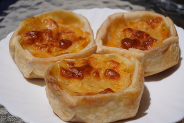 Close-up of three small baked pastries with golden, crispy cheese filling on a white plate. The flaky dough and browned tops highlight a homemade, warm and appetizing savory snack.