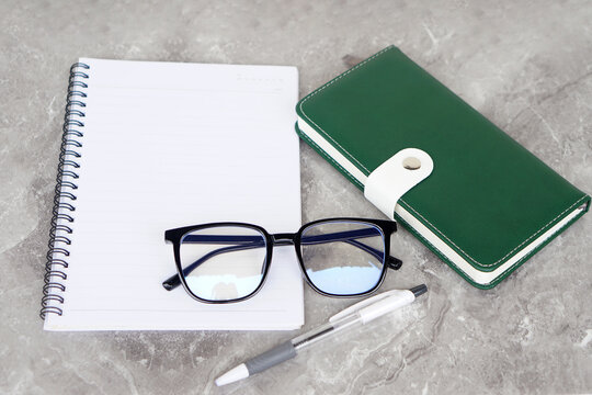 A flat lay composition featuring an open spiral notebook, a pair of eyeglasses, a green planner with a white clasp, and a pen on a gray marble surface, symbolizing organization and productivity.