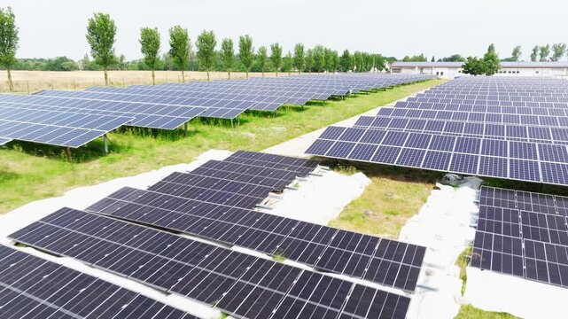 The drone flies over a solar field from a top-down view. Evenly arranged solar panels create a geometric pattern across the green area.