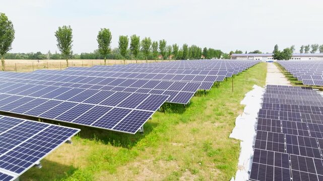 The drone flies over a solar field from a top-down view. Evenly arranged solar panels create a geometric pattern across the green area.