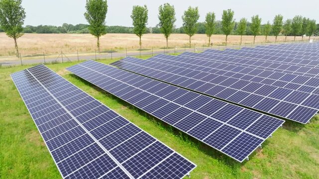 The drone flies over a solar field from a top-down view. Evenly arranged solar panels create a geometric pattern across the green area.