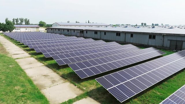 The drone flies over a solar field from a top-down view. Evenly arranged solar panels create a geometric pattern across the green area.