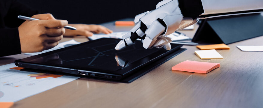 A human hand collaborates with a robotic hand on a touchscreen tablet in a modern office setting filled with colorful sticky notes and documents, symbolizing innovation.Noogenesis
