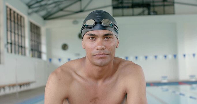 Leaning shirtless swimmer preparing at pool edge in pool hall, wearing black swim cap and goggles