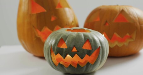 Fototapeta premium Glowing green-gray carved pumpkin sitting on white tabletop in studio, with two lit orange pumpkins