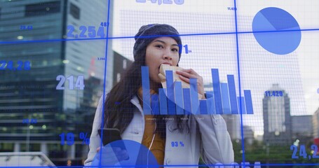 Biting sandwich, woman in knit beanie and coat checking smartphone in plaza, with finance charts