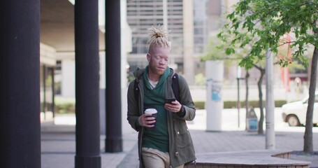 Walking man glancing at phone while holding coffee in plaza, wearing green hoodie with backpack