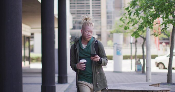 Walking man glancing at phone while holding coffee in plaza, wearing green hoodie with backpack