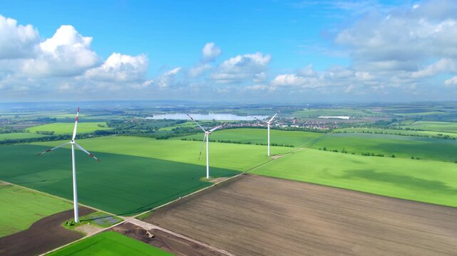 The drone glides smoothly over wide fields and several wind turbines. Clear visibility, green landscapes, and soft summer light define the scene.