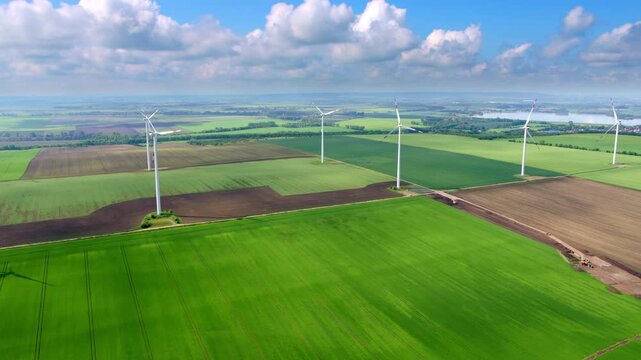 The drone glides smoothly over wide fields and several wind turbines. Clear visibility, green landscapes, and soft summer light define the scene.