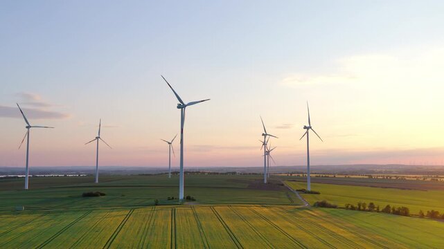 The drone flies smoothly over a wind farm, capturing several turbines in a wide green landscape during soft evening light.