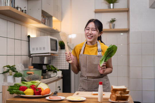 Happy Asian woman holding a wooden spoon and lettuce, ready to cook a healthy meal in her kitchen