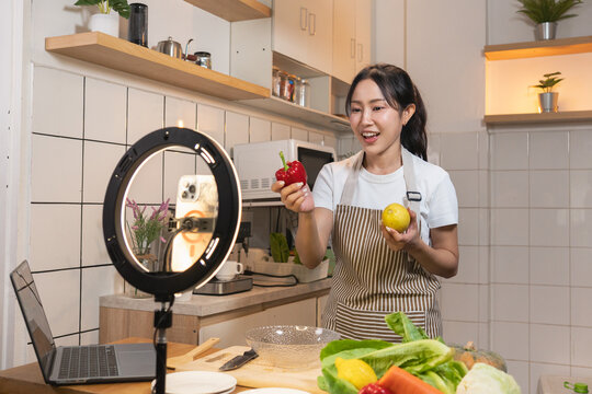 Happy Asian food vlogger talking and showing ingredients to her audience during a live stream cooking session