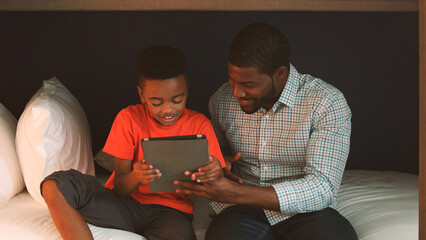 Father With Son Sitting In Bedroom Playing Game On Digital Tablet Together