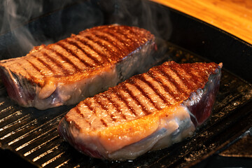 Two Grilled Beef Steaks with Grill Marks Sizzling on a Hot Cast Iron Pan. Juicy, seared meat with a delicious crispy crust and visible smoke. Perfect for BBQ concepts.