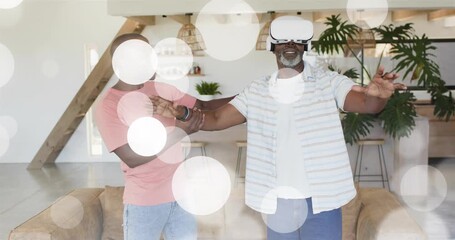 Senior wearing VR headset reaching forward, son steadying arm, bokeh overlays aiding tech care - Powered by Adobe