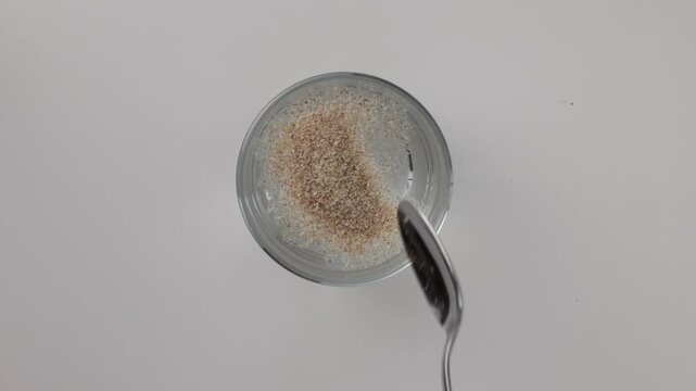 Pouring psyllium into a glass of water and stirring with a silver spoon, top view on a white background