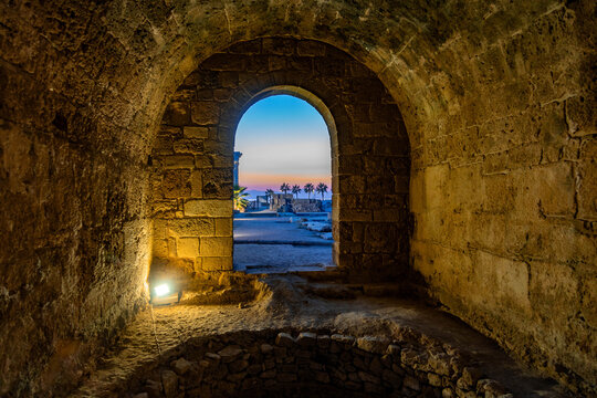 Byzantine stone vault with archway framing view of Apollo Temple and palm trees against orange sunset sky at ancient archaeological site. Side, Antalya, Turkey.

