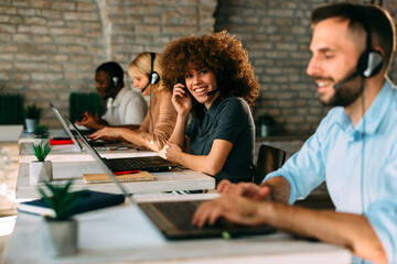 Smiling female customer service agent or call center employee sits at her desk with a headset, working enthusiastically in a modern, diverse office with her colleagues