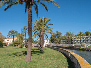 Fototapeta premium Tropical garden scene featuring palm trees, a grass lawn, a winding canal, and resort buildings under a clear sky.