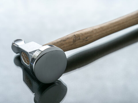 Close-up of a highly polished jewelry chasing hammer with a wooden handle on a reflective, gray surface.