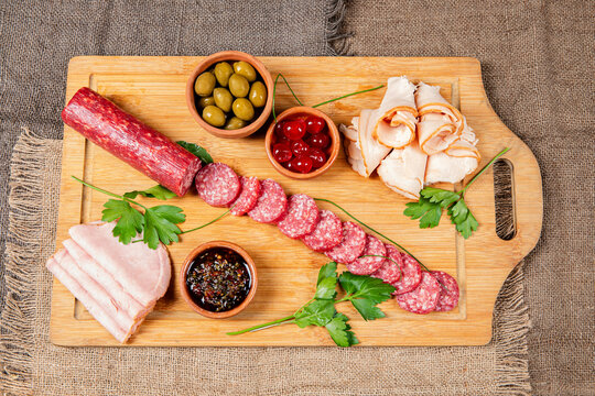 Rustic board with cold cuts, olives, cherries, and sauce on burlap. Ideal for food presentations, events, or premium culinary content.